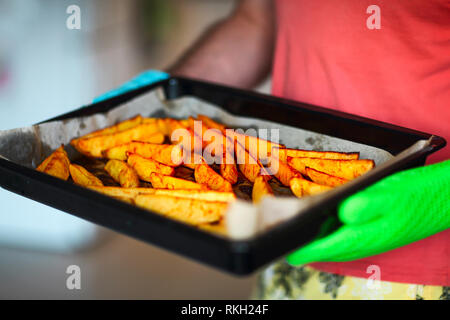 Close up shot of roast potatoes and parsnips in a tray. A man is holding it in his hands wearing oven gloves. Party time Stock Photo