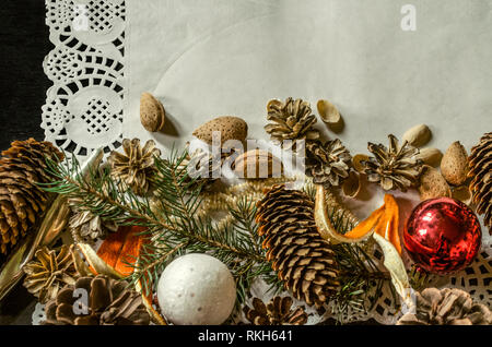 The bottom edge of the openwork paper napkin with various pine cones, fir branch, old worn Christmas toys, almonds and orange peel on a black backgrou Stock Photo