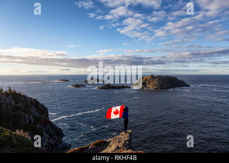 Adventurous woman holding a Canadian Flag on a Rocky Atlantic Ocean Coast during a sunny day. Taken in Sleepy Cove, Crow Head, Twillingate, Newfoundla Stock Photo