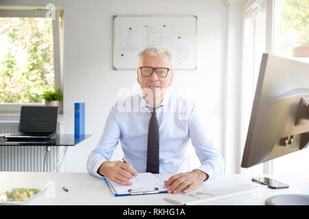 Portrait of elderly businessman sitting at office desk and doing some paperwork while looking at camera and smiling. Stock Photo