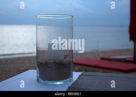 Glass container with white candle and sand as table decoration for romantic dinner on the seaside during sunset Stock Photo