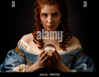 Lovely Woman in elegant medieval dress holding small glass ball and looking at camera while standing on black background Stock Photo
