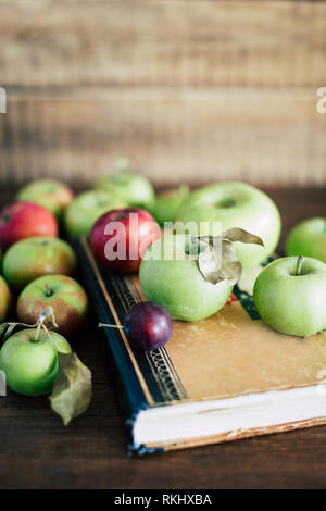 Bunch of ripe apples and small plum lying on shabby old book on wooden tabletop Stock Photo
