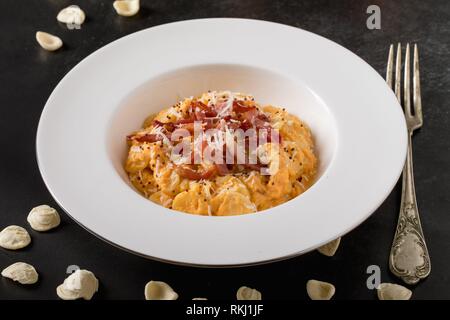 Close-up of plate with orecchiette pasta garnished with pumpkin cream, roasted bacon and parmesan cheese, on black background.