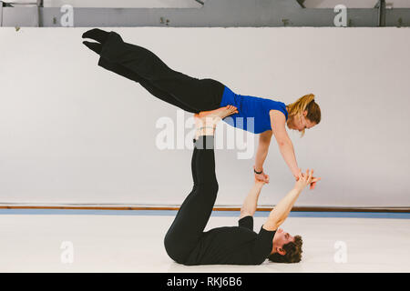 Theme is sports and acreage. A young Caucasian male and female couple practicing acrobatic yoga in a white gym on mats. a man lies on his back and hol Stock Photo