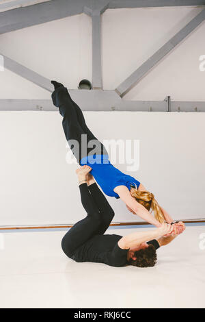 Theme is sports and acreage. A young Caucasian male and female couple practicing acrobatic yoga in a white gym on mats. a man lies on his back and hol Stock Photo