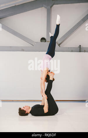 Theme is sports and acreage. A young Caucasian male and female couple practicing acrobatic yoga in a white gym on mats. a man lies on his back and hol Stock Photo