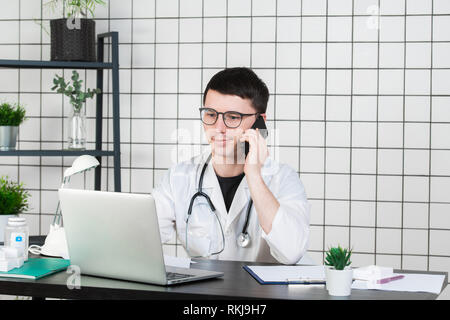 Male doctor using telephone while working on computer at table in clinic Stock Photo