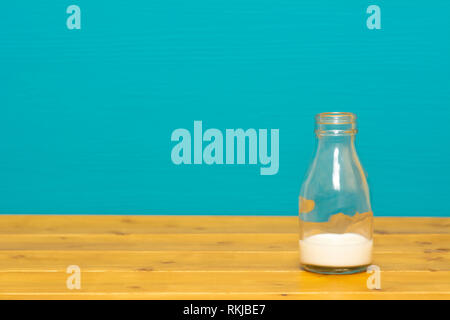 One-third pint glass milk bottle with dregs of fresh creamy milk, on a wooden table against a bright teal painted background Stock Photo