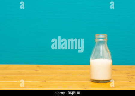One-third pint glass milk bottle half full with fresh creamy milk, on a wooden table against a bright teal painted background Stock Photo