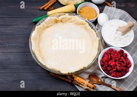 Pie base in a glass round form and Ingredients for making cake  - flour, egg, cherry, brown sugar, cinamon on the dark old wooden table. Making cherry Stock Photo