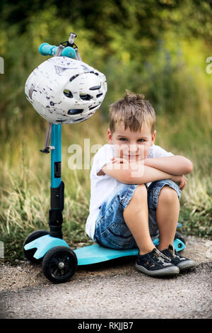 A small boy with a helmet sitting on scooter on a road in park on a summer day. Stock Photo