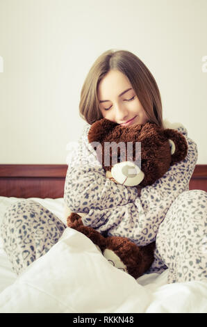 Happy young woman with blonde hair sitting on the bed in her bedroom in an embrace with a stuffed animal toy. Favorite brown teddy bear in the hands o Stock Photo