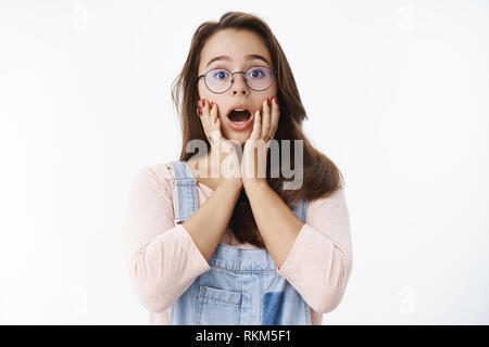 Waist-up shot of surprised and amazed young cute female geek in glasses dropping jaw saying wow holding hands on cheeks impressed and astonished with Stock Photo