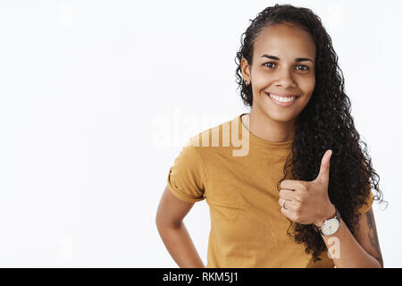 Nice work, keep up. Satisfied happy ougoing dark-skinned girl with curly hair showing thumbs-up gesture and smiling broadly, liking great effort of Stock Photo