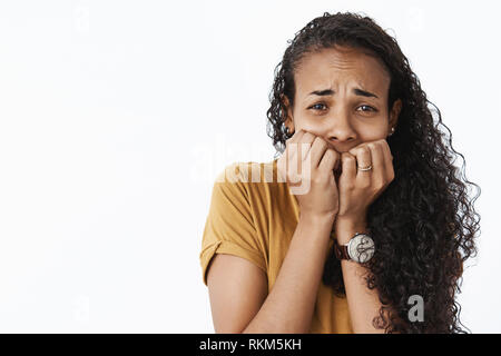 Close-up shot of terrified scared timid african american woman frowning trembing from fear as seeing spooky movie scene, holding fingernails on mouth Stock Photo
