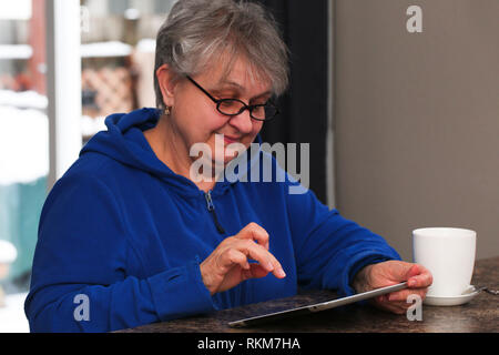 Beautiful, Mature, Elder, Grandmother, Woman with Grey Hair, in Eyeglasses is Using a Digital Tablet, Smiling, at home Stock Photo