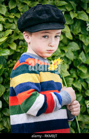 Sad, cute little boy in rainbow multicolored striped sweater and black suede newsboy cap holding an orange daisy flower Stock Photo