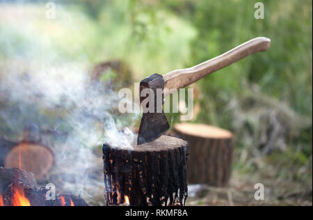 Axe in tree stump and campfire with smoke in summer forest at sunny day Stock Photo