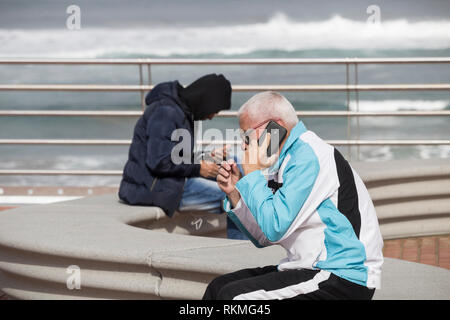 Elderly man and young man sitting on seat oveloooking the sea. Both using mobile phones Stock Photo