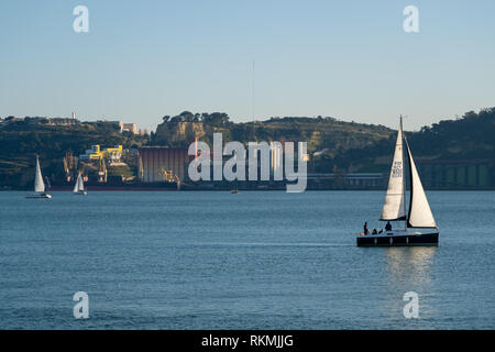 Lisbon, Portugal - 12/28/18: Sailboats on tagus river, sun shining on white sails, calm waters with big factory buildings in the background riverside Stock Photo