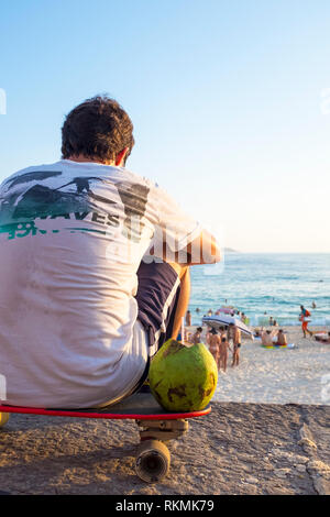 RIO DE JANEIRO - FEBRUARY 08, 2015: A young Brazilian man sits with a green drinking coconut on his skateboard overlooking Ipanema Beach at Arpoador. Stock Photo