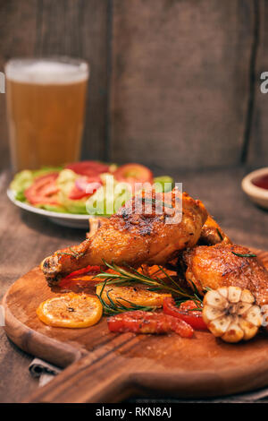 Grilled chicken legs roasted on the grill on wooden chopping board with tomato sauce in a bowl, fresh tomatoes and lettuce leaves, bitter pepper, glas Stock Photo