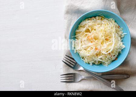 Blue bowl of shredded chopped pickled cabbage and carrot. Served with two forks on folded grey fabric towel and viewed from above on white table surfa Stock Photo
