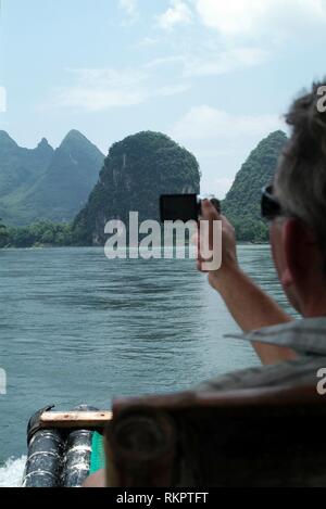 A tourist photographs the spectacular scenery along the Li River while floating on a bamboo raft in Yangshuo. Speckled with karst mountains and the ge Stock Photo