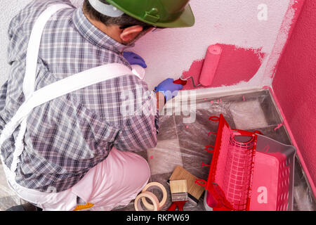 Caucasian house painter worker in white work overalls, with the roller he paints the wall with the colored painting of pink. Construction industry. To Stock Photo