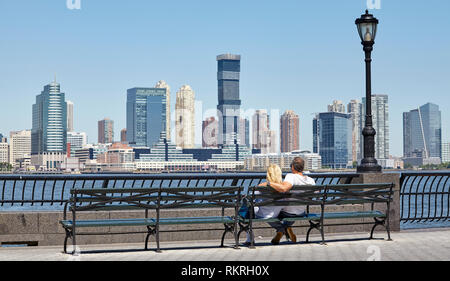 New York, USA - July 08, 2018: A couple sits on a bench at Hudson River promenade with view of Jersey City on a sunny summer morning.