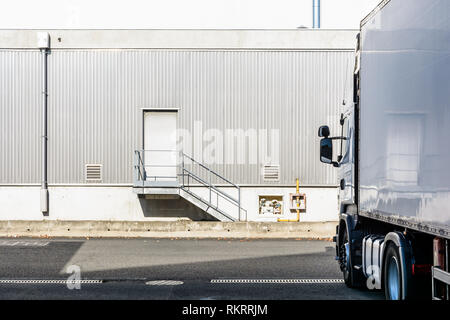 A semitrailer truck parked in front of an industrial building covered with a grey corrugated iron siding, with a door at the top of a small staircase. Stock Photo