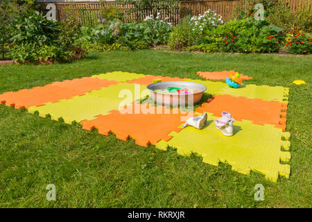 Color children's mat on the green grass in the country. Porous orange foam mat carpet. Children's shoes in the foreground Stock Photo