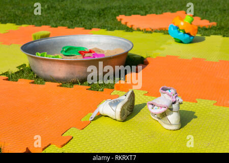 Color children's mat on the green grass in the country close-up. Handmade sandpit. Porous orange foam mat carpet. Children's shoes in the foreground.  Stock Photo