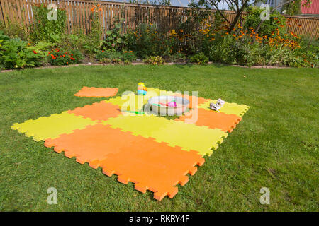 Color children's mat on the green grass in the country close-up. Handmade sandpit. Porous orange foam mat carpet. Children's shoes in the foreground.  Stock Photo