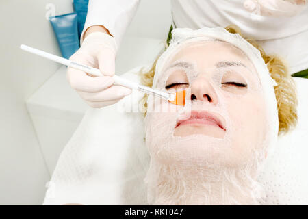 beautician worker holding paintbrush  applying facial cosmetic serum  in biological treatment to female client  in beauty salon, eyes closed, close up Stock Photo
