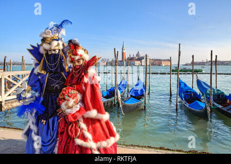 Two participants wear red and blue dresses, costumes and masks standing near gondolas on Grand Canal and San Giorgio Maggiore church in Venice. Stock Photo