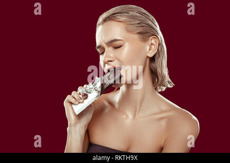 Blonde-haired model biting chocolate bar standing near red wall Stock Photo