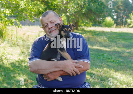 Outdoor portrait of Caucasian senior man relaxing while holding black little puppy on the hands Stock Photo