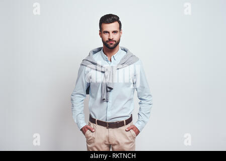 Irresistible man. Bearded young man is looking at camera, putting his hands in his pockets while standing against grey background. Beauty look. Close up Stock Photo