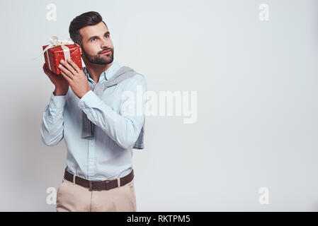 What is inside? Attractive stylish man is holding a gift box near his ear and trying to guess what is inside while standing on a grey background. Close-up image Stock Photo