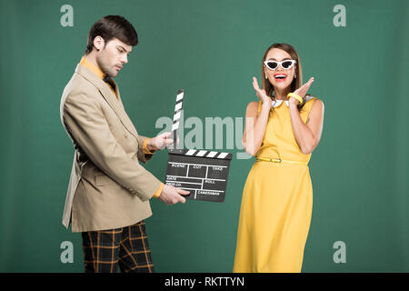 handsome man in vintage clothes holding film clapperboard while happy woman gesturing with hands isolated on green Stock Photo