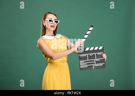 beautiful stylish woman in yellow dress and sunglasses holding film clapperboard isolated on green Stock Photo
