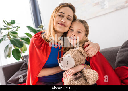 Hugging mother and kid in red cloaks holding teddy bear Stock Photo