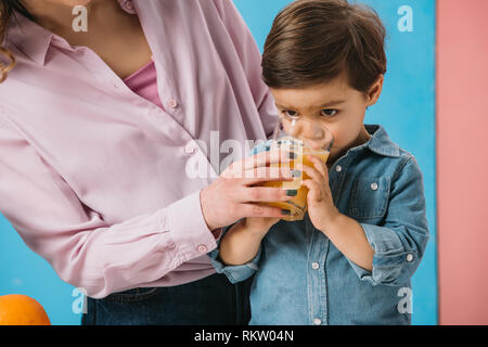 cute little boy drinking fresh orange juice while holding glass with mothers hand on bicolor background Stock Photo