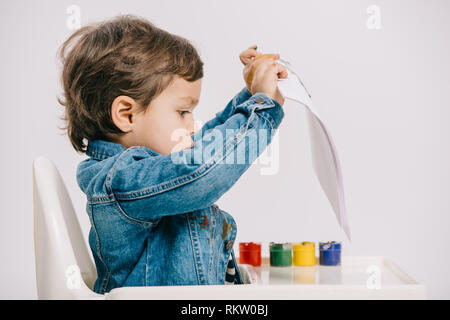 cute little boy holding paper while sitting on highchair with watercolor paints on table isolated on white Stock Photo