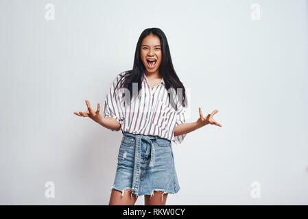 I did it. Happy excited asian woman with dark hair in casual wear keeping arms raised and looking at camera while standing against grey background. Success concept. Close-up portrait. Stock Photo