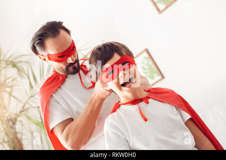 Father and little son wearing superheroe costumes together at home standing man tying knot on mask of boy looking camera laughing cheerful Stock Photo