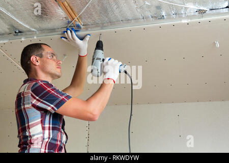 Young man in goggles fixing drywall suspended ceiling to metal frame using electrical screwdriver on ceiling insulated with shiny aluminum foil. Renov Stock Photo