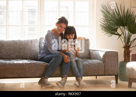 Happy mom and kid daughter using smartphone sitting on couch Stock Photo